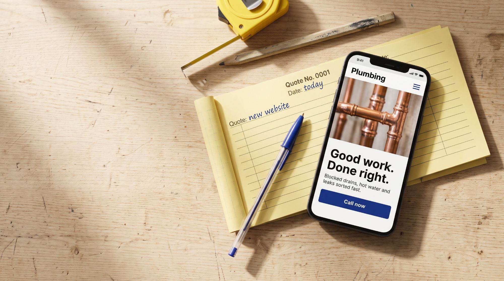 Phone showing an example plumbing website on a timber workbench beside a yellow quote docket and biro